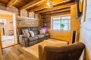 L’intérieur d’un chalet confortable, avec un canapé en cuir, un petit fauteuil et une fenêtre donnant sur un paysage verdoyant.