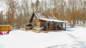 Une cabane en rondins dont le toit est recouvert de neige, avec une véranda dotée de chaises et d’un hamac.