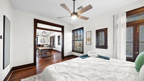 Une chambre dotée d’un grand lit, d’un plancher en bois, d’un ventilateur de plafond et offrant une vue sur un coin repas grâce à une porte ouverte.