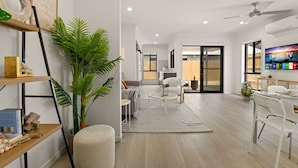 Una sala de estar moderna con sofá, mesa de centro y televisor. Hay una planta, una estantería y un comedor con sillas.