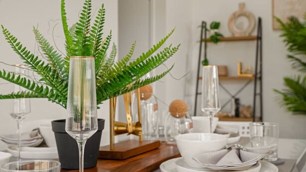 Una mesa de comedor dispuesta con platos y cuencos blancos, y un centro de mesa con un helecho verde en un florero de vidrio.