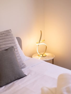 A neatly made bed with a white sheet and a gray pillow, a small round table with a modern lamp, and a plain wall in the background.