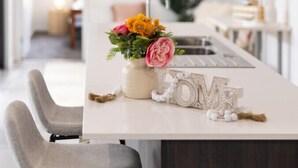 Eine moderne Küche mit weißer Arbeitsplatte, zwei grauen Stühlen, einer Blumenvase und Dekorationselementen, darunter ein Traumfänger und ein Schild mit dem Wort 'LOVE'.