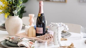 Ein Esstisch gedeckt mit Tellern, einer Champagnerflasche, einer Vase mit gelben Blumen und einer dekorativen Schale.