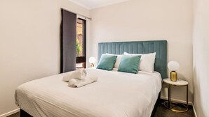 Un lit bien fait, recouvert d’une couette blanche et agrémenté d’oreillers turquoise, une table de chevet ronde à cadre doré avec une lampe blanche, et une fenêtre avec un rideau foncé.
