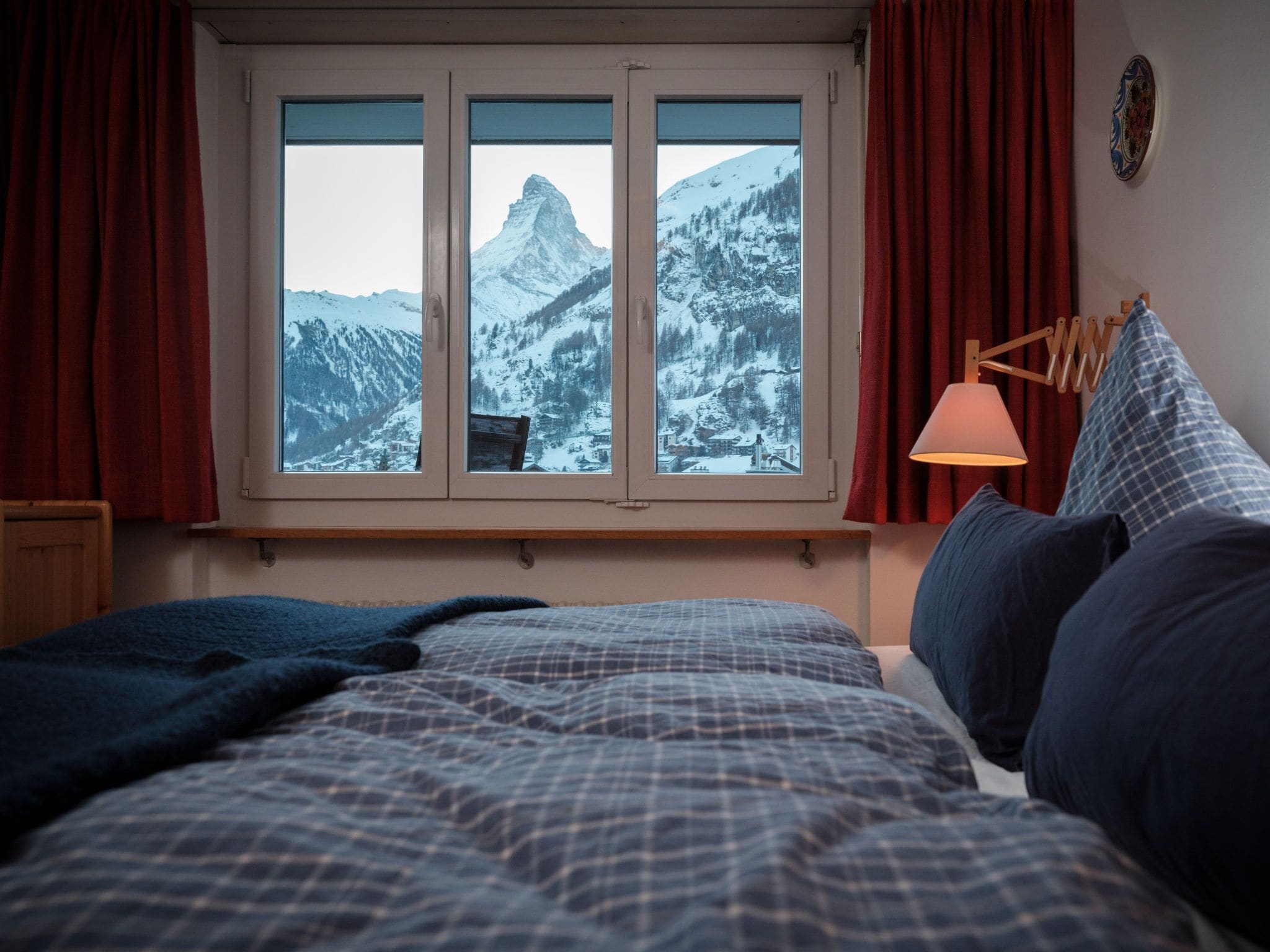 A bedroom with a bed, a window with a view of snow-covered mountains, and a bedside lamp.