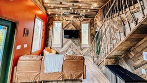 Interior rústico de una cabaña con techo de madera, un sofá, un televisor y una escalera.