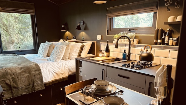 Un dormitorio compacto y bien iluminado con cama, mesita de noche y ventana. Junto a él, una kitchenette con estufa, lavamanos y diversos utensilios de cocina.