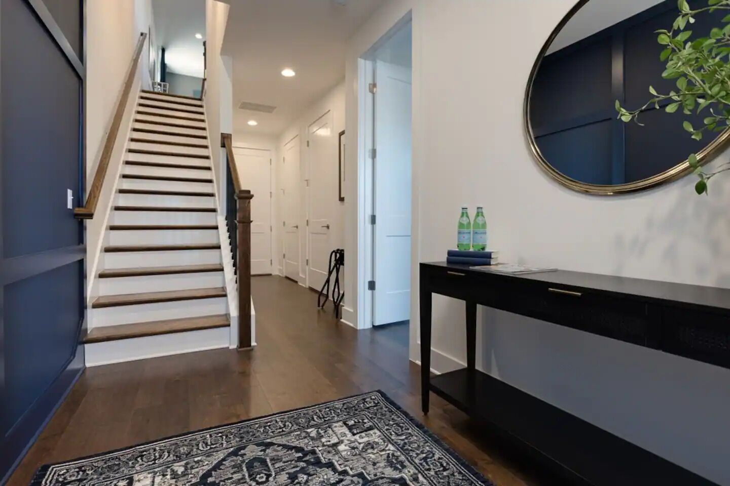 Un couloir avec un escalier en bois, une console noire sur laquelle sont posées deux bouteilles d’eau, et un tapis à motifs.