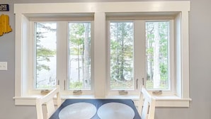 A dining area with a table and chairs, large windows with a view of trees and a body of water, and a sign on the wall. - Quiet Leonard Lake Retreat,  Beautiful Waterfront Home  (Ellsworth)