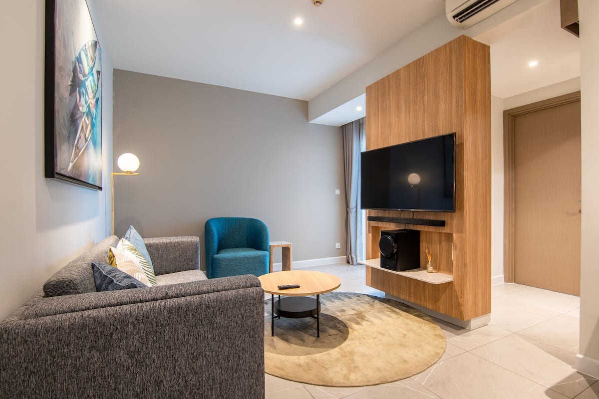 Ein modernes Wohnzimmer mit grauem Sofa, blauem Sessel, rundem Couchtisch und einem Flachbildfernseher an einer Holzwand.