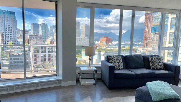 A modern living room with a large window offering a city view, a dark-colored sofa with patterned cushions, and a small side table with a lamp. - Amazing Views, DT, Free Parking  (Vancouver)