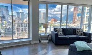 A modern living room with a large window offering a city view, a dark-colored sofa with patterned cushions, and a small side table with a lamp. - Amazing Views, DT, Free Parking  (Vancouver)