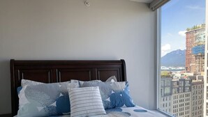 A bed with a patterned bedspread and multiple pillows, a wooden headboard, and a large window with a cityscape view. - Amazing Views, DT, Free Parking  (Vancouver)