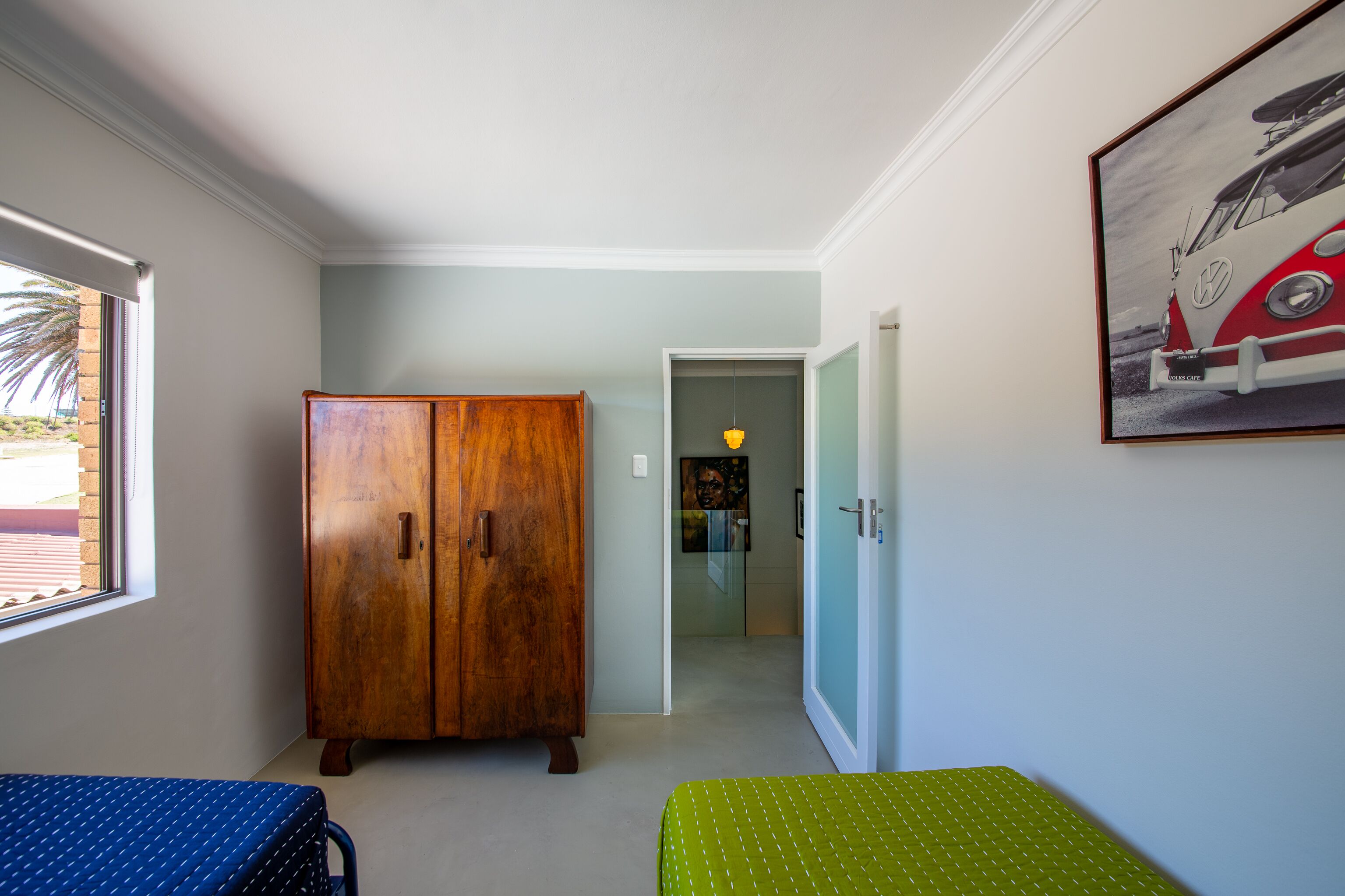 Een slaapkamer met een houten garderobe, een bed met een deken met een blauw en groen patroon, en een lijst met daarop een foto van een oldtimer.