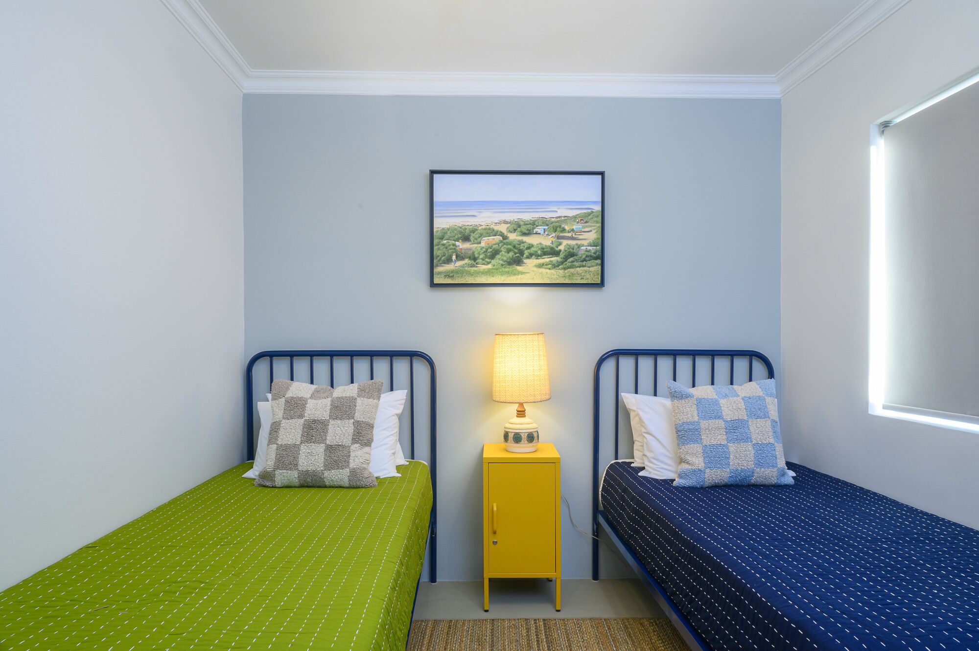 A room with two single beds, a small yellow cabinet, a wall-mounted lamp, and a framed landscape painting.