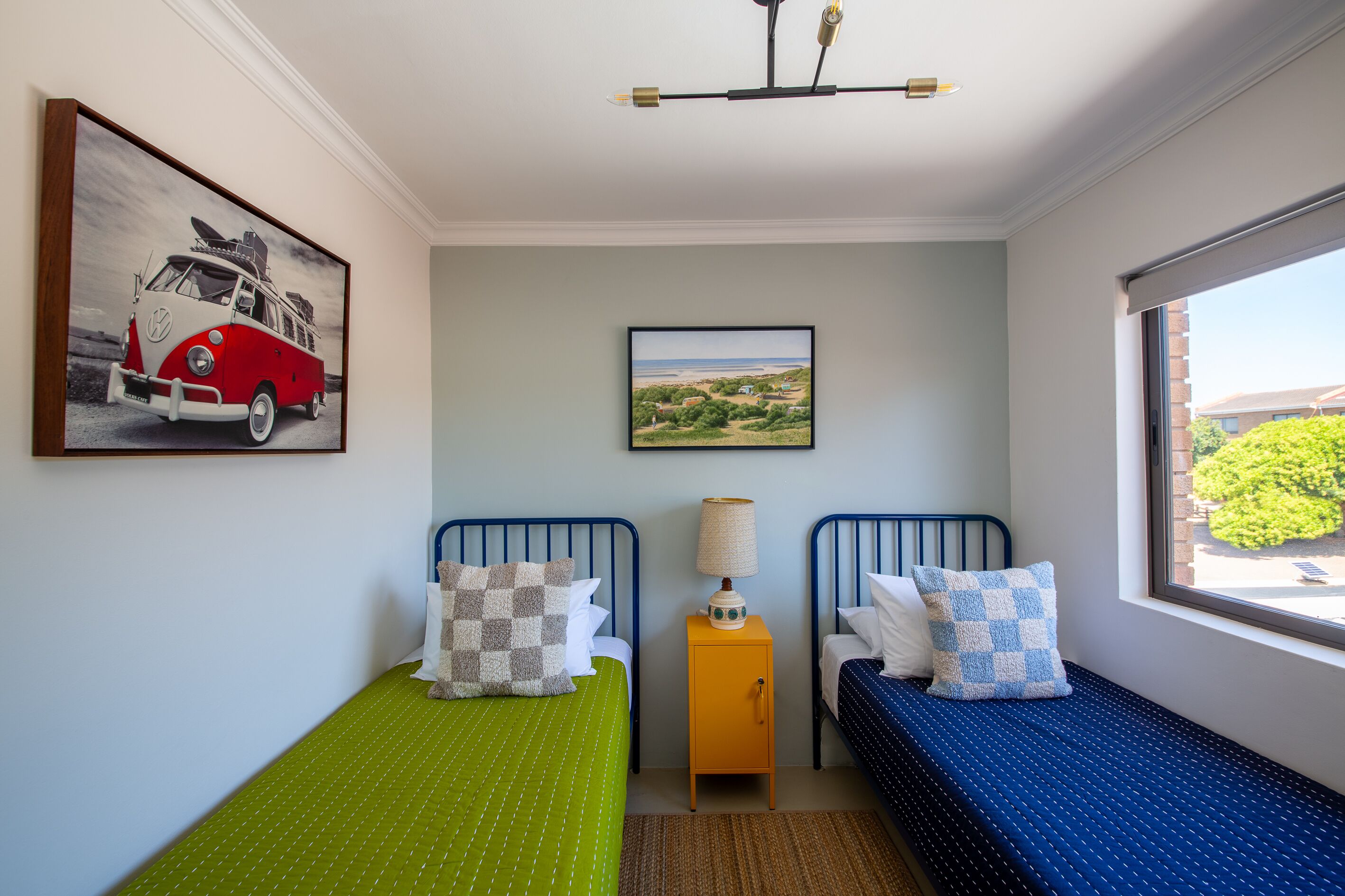 Une chambre avec deux lits, une fenêtre donnant sur un beau paysage, un tableau représentant une voiture et une lampe sur une table d’appoint.