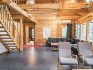 A wooden interior with a staircase, a living area with a sofa and chairs, and large windows.
