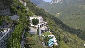 A large, white villa with multiple balconies, surrounded by lush greenery and mountains.