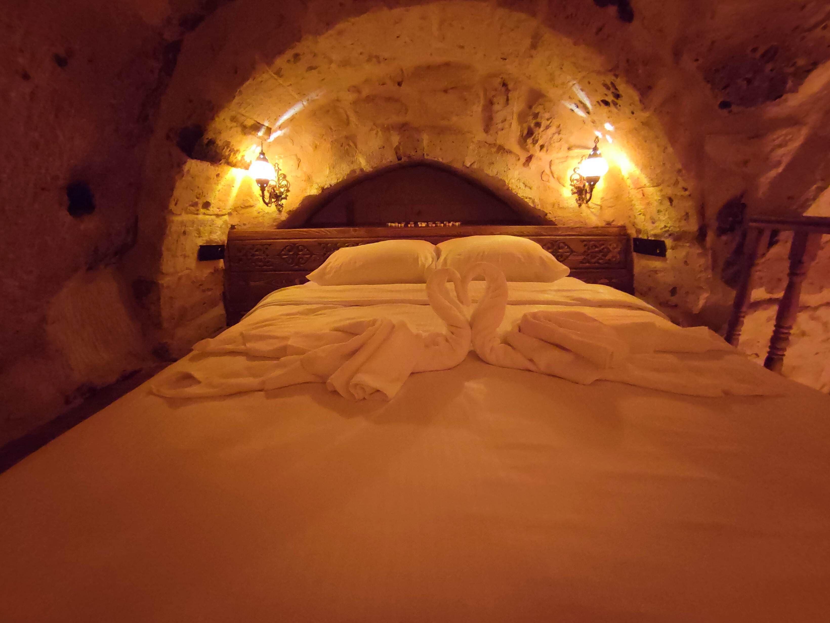 Una cama con sábanas blancas y dos toallas con forma de cisne en una habitación de paredes de piedra con dos luces fijadas a la pared.