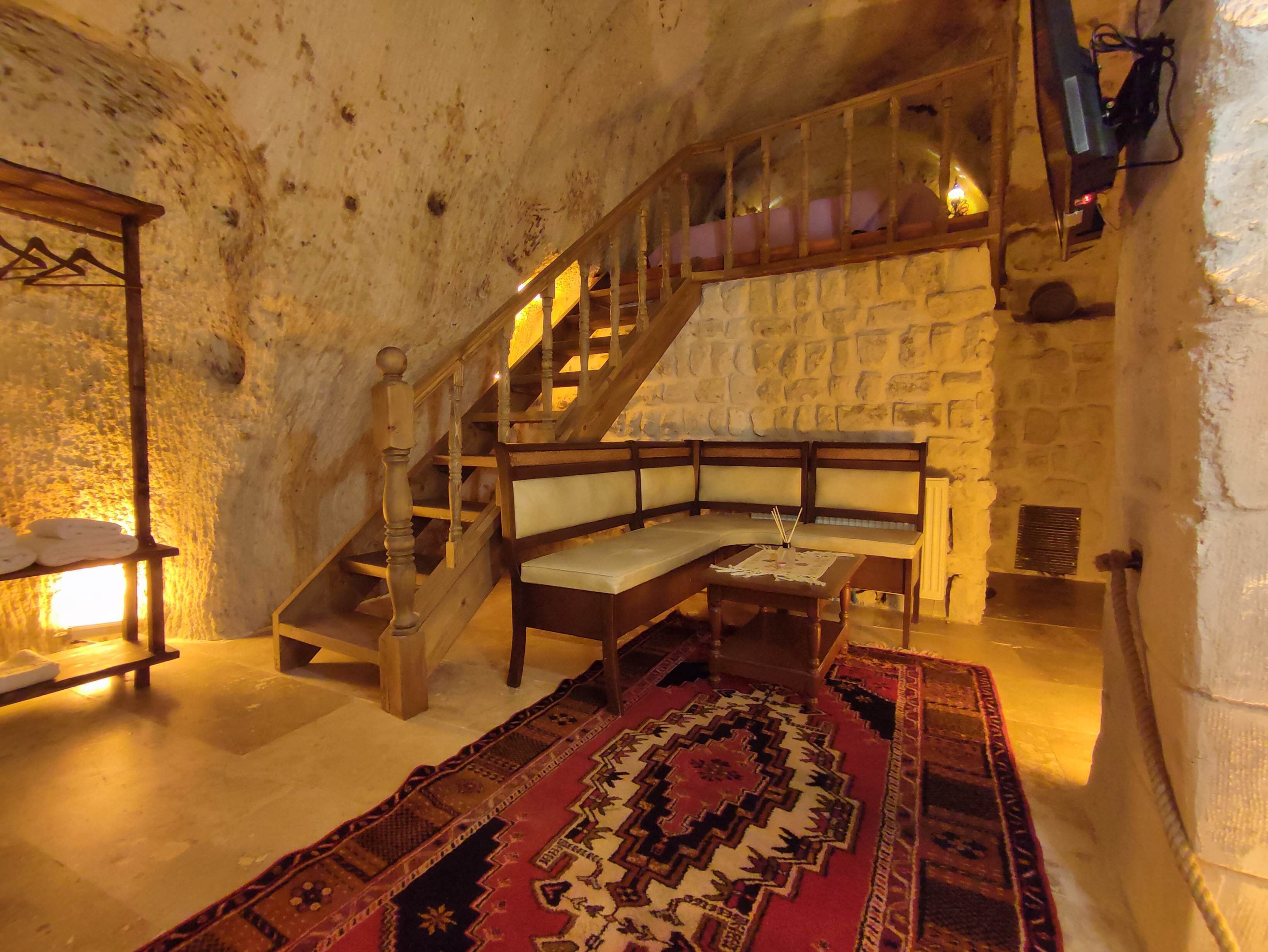 A rustic room with a stone wall, a wooden staircase, a bench, a patterned rug, and a small table with a candle.