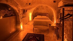 A cozy, dimly lit room with a bed, a small table, and a bench. The walls are textured and appear to be made of stone or a similar material. - Euphoria Cave House (Nevsehir)