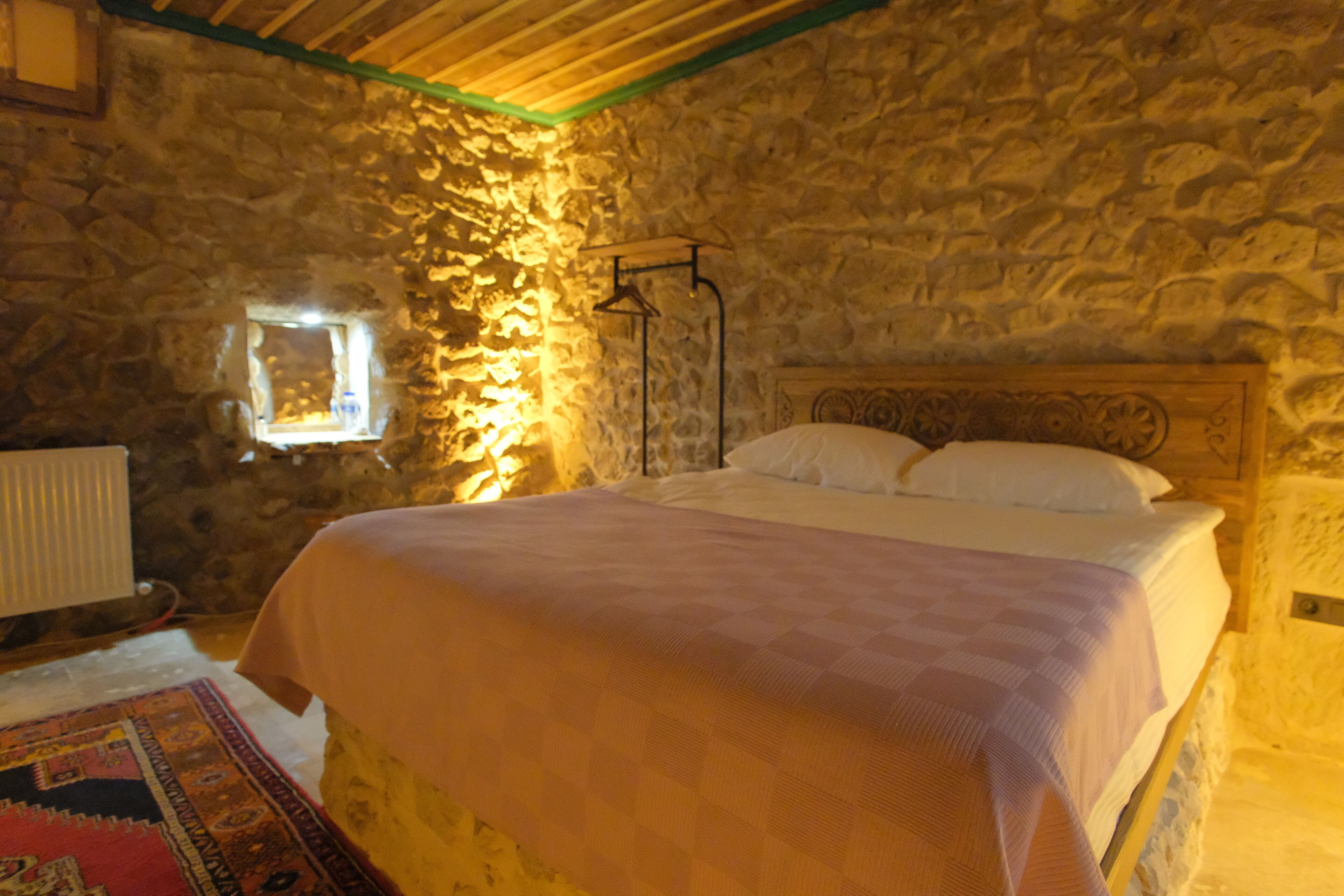 A bedroom with a stone wall, a wooden headboard, a bed with white linens, and a small window with a light fixture.