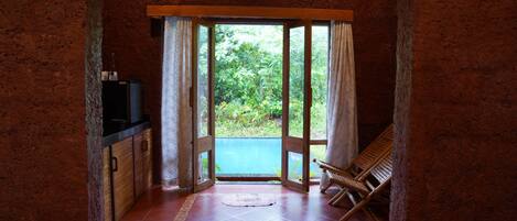 A room with a kitchen area, a wooden chair, and a view of greenery through a glass door.