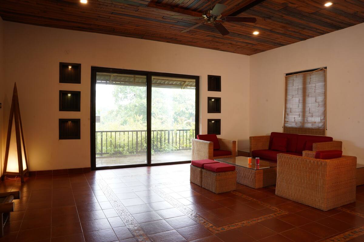 A spacious living area with a wooden ceiling, a large sliding glass door leading to a balcony, and wicker furniture with red cushions.