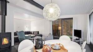 A modern living room with a fireplace, a dining table with a tray of bread and cups, and a large window with a view of trees.