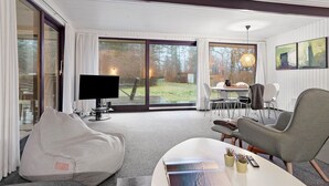 A modern living room with a large TV, comfortable seating, a dining area, and a view of the outdoors.