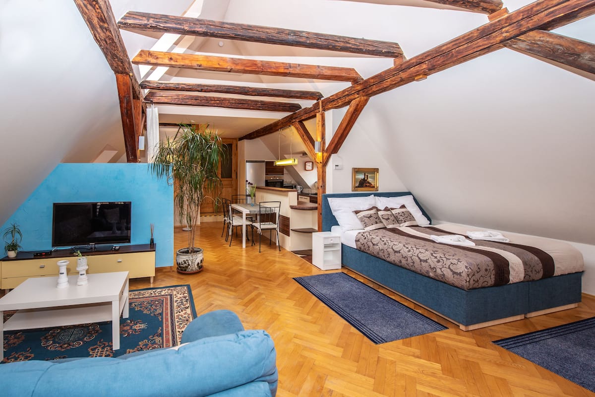 A loft-style bedroom with exposed wooden beams, a bed, a desk, and a television.