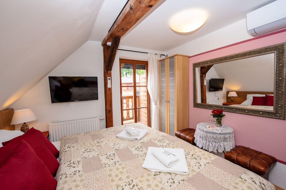 A bedroom with a large bed, a television, a mirror, a small table with flowers, and a window with curtains.