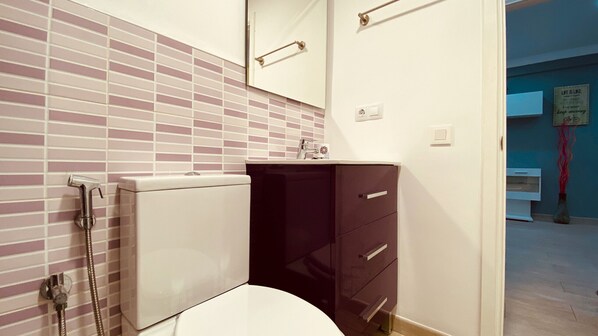 Ein Badezimmer mit Toilette, Waschbecken und Spiegel. Darüber hängt ein Handtuchhalter und an der Wand ist ein Duschkopf.