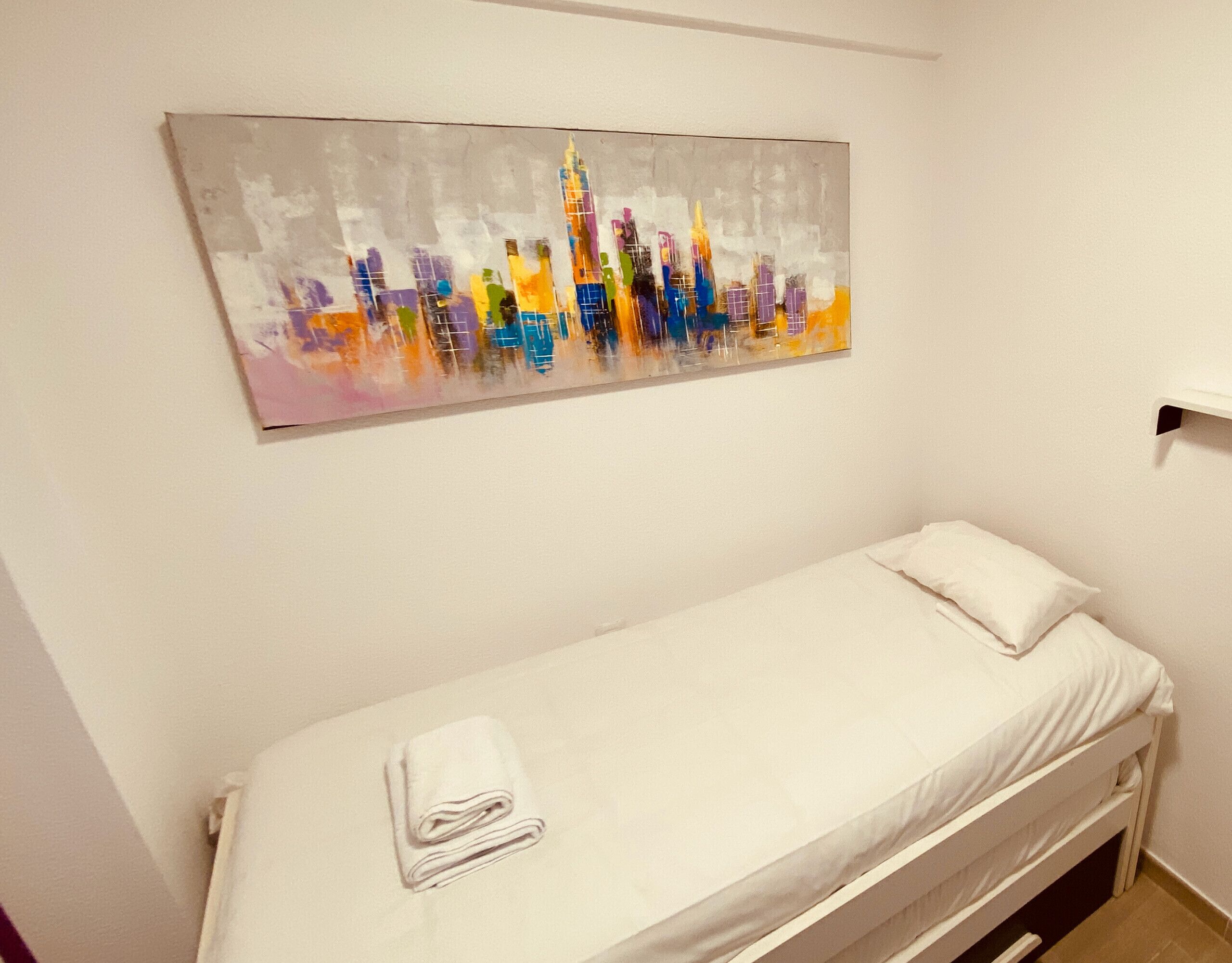 A single bed with a white mattress and pillow, a folded white towel, and a colorful abstract painting on the wall.