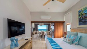 A modern hotel room with a large flat-screen TV, a bed with patterned bedding, a wooden console table, and a view of the ocean through the window. - Soleado 312 Oceanfront Condo at Costa Azul Beach! (San José del Cabo)