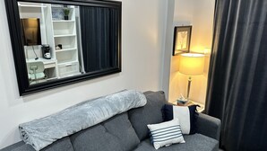 Una sala de estar moderna con un sofá gris, un espejo grande, una mesita de noche con una lámpara y una cortina oscura.