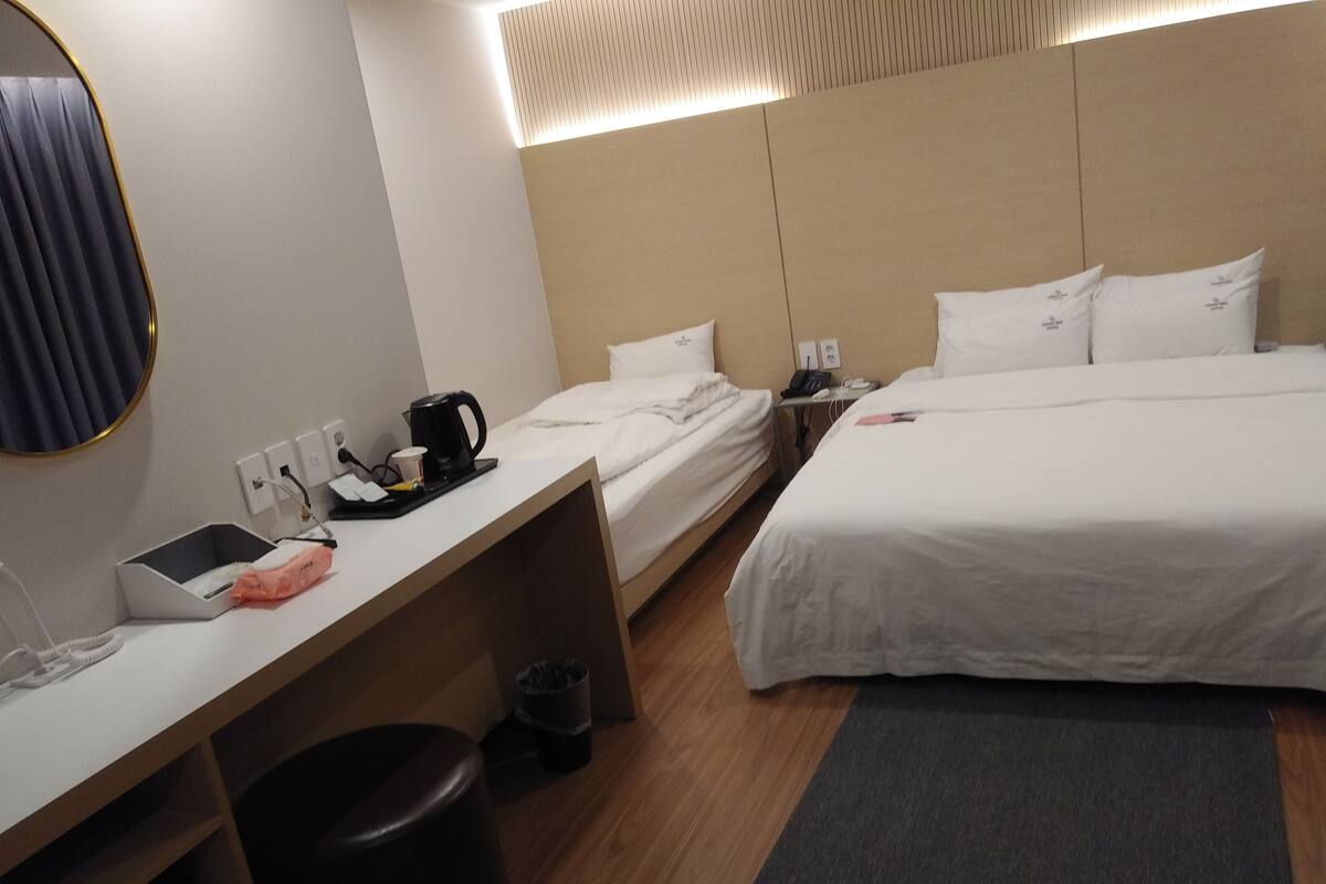 A hotel room with two beds, a desk with a coffee maker, a trash bin, and a small table with a phone and a lamp.