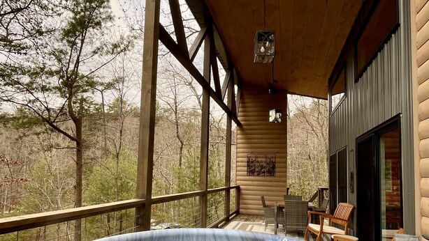 A hot tub on a wooden deck with a view of trees, a covered seating area, and a large window. - Whispering Woods • Modern Cabin w/ HOT TUB (Stanton)