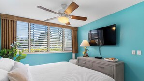 A bedroom with a bed, a ceiling fan, a TV, a window with blinds, and a nightstand with a lamp. - Oceanfront Beachfront Escape Hale Kai 1Bed (Lahaina)