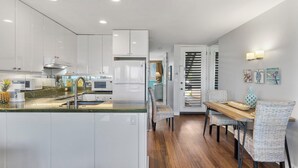 A modern kitchen with white cabinets, a central island, and a dining area with wicker chairs and a wooden table. - Oceanfront Beachfront Escape Hale Kai 1Bed (Lahaina)