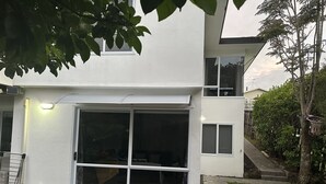 Una casa moderna de dos pisos con un amplio acceso por puerta corredera de vidrio, un espacio exterior visible con una silla de metal y un aro de baloncesto en un árbol.