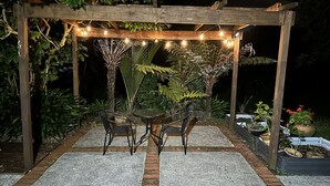 Una pérgola de madera con luces colgantes, dos mesas con sillas y plantas en macetas.