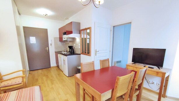 A compact kitchen with a refrigerator, stove, and microwave. There is a dining table with chairs and a television on a stand. - Appartement T2 Centre Luchon-wifi - Vacances et Cure - à 280m de la Télécabine (Bagnères-de-Luchon)