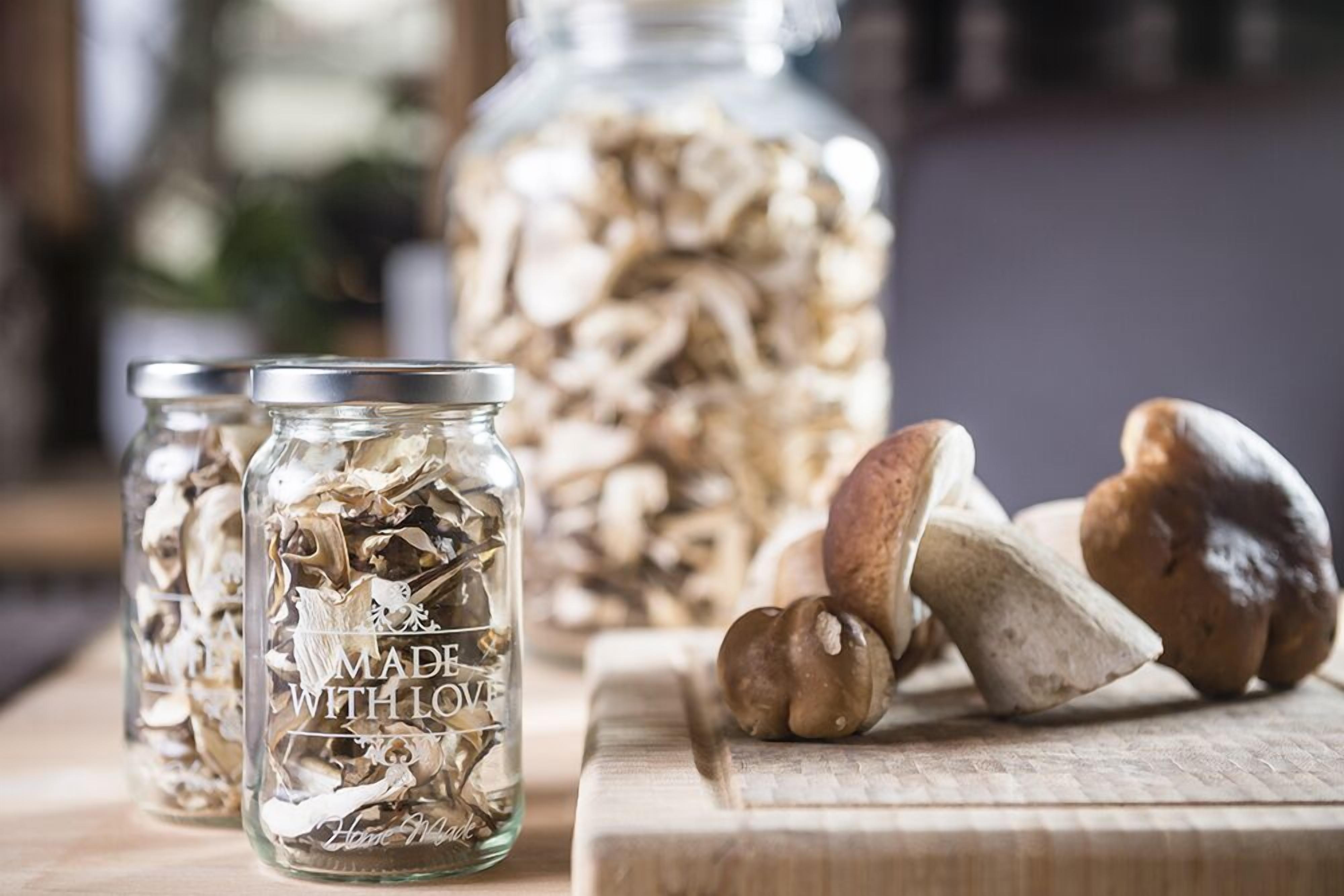 Three glass jars with dried mushrooms on a wooden surface, with a larger jar in the background.