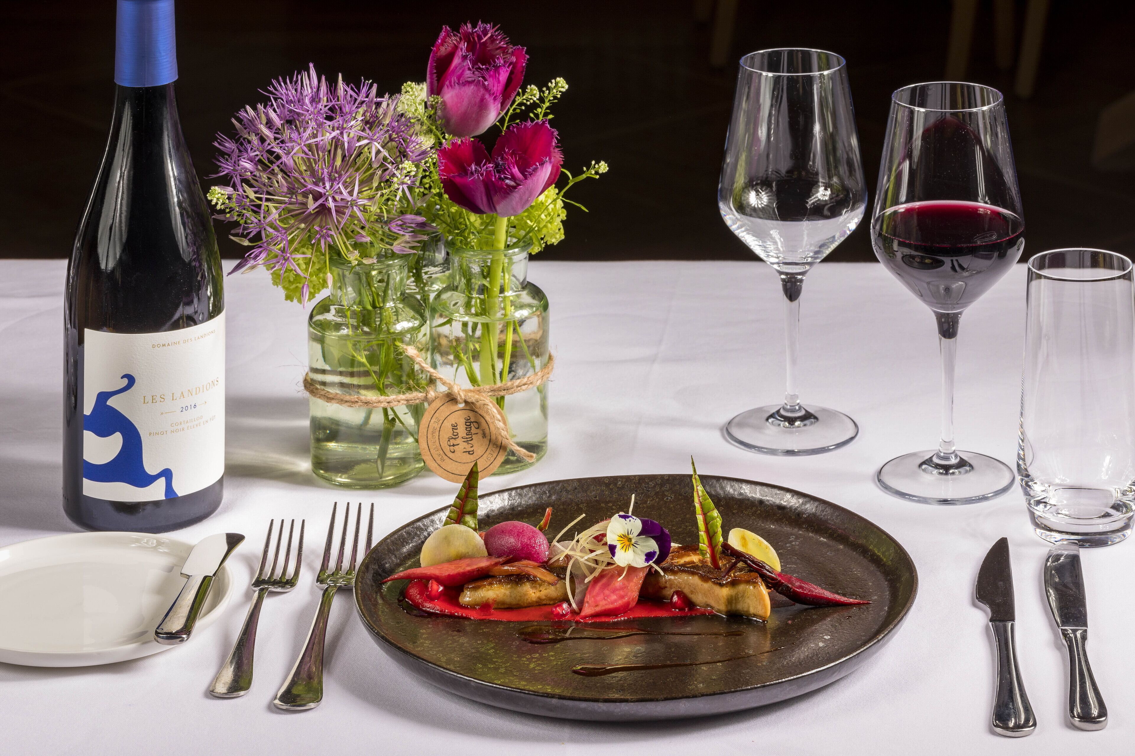 Una mesa de comedor dispuesta con una botella de vino, un florero con flores y un plato de comida.