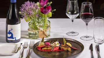 A dining table set with a bottle of wine, a vase with flowers, and a plate of food.