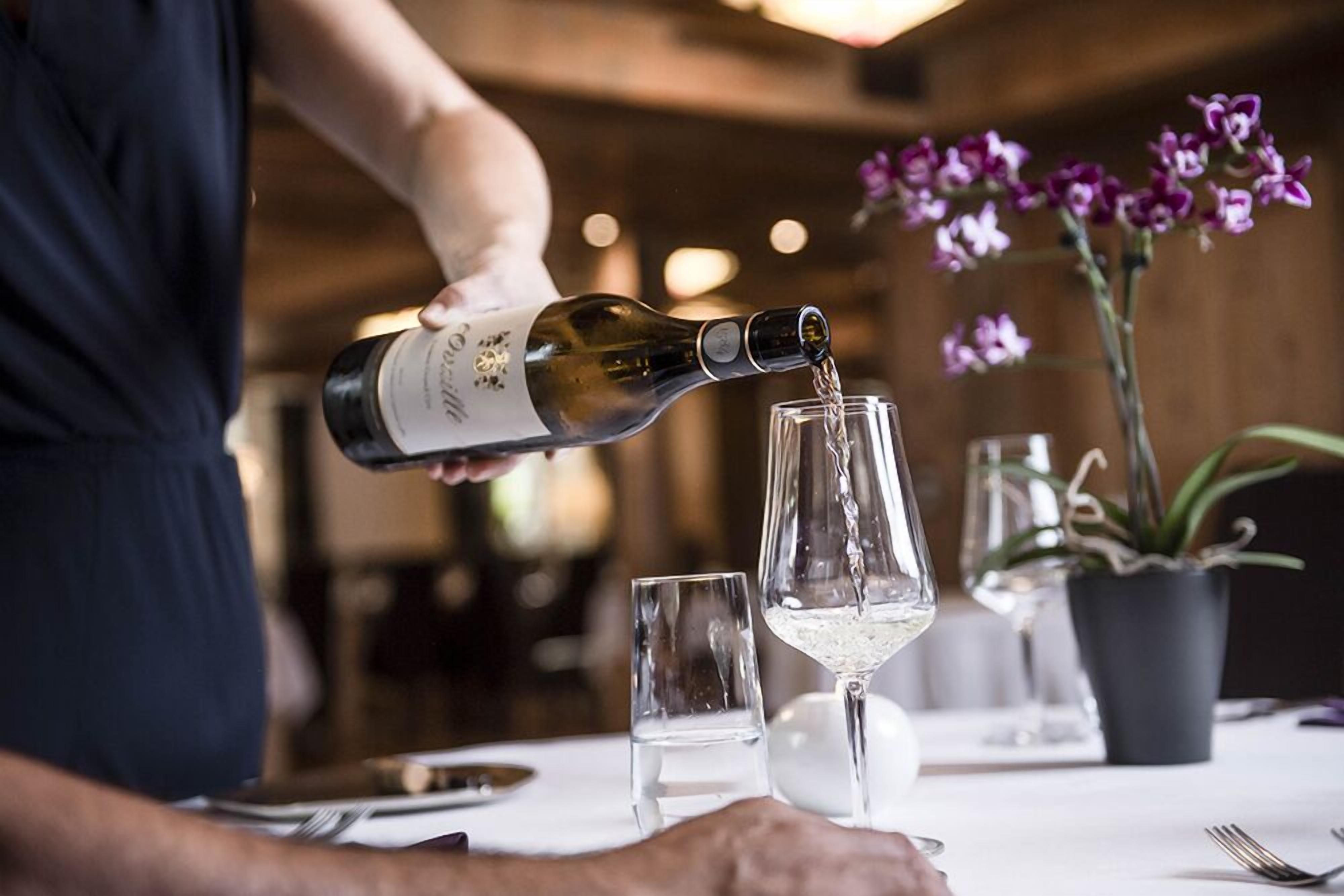 Una persona sirviendo vino en una copa sobre una mesa con una orquídea en maceta y un pequeño florero.