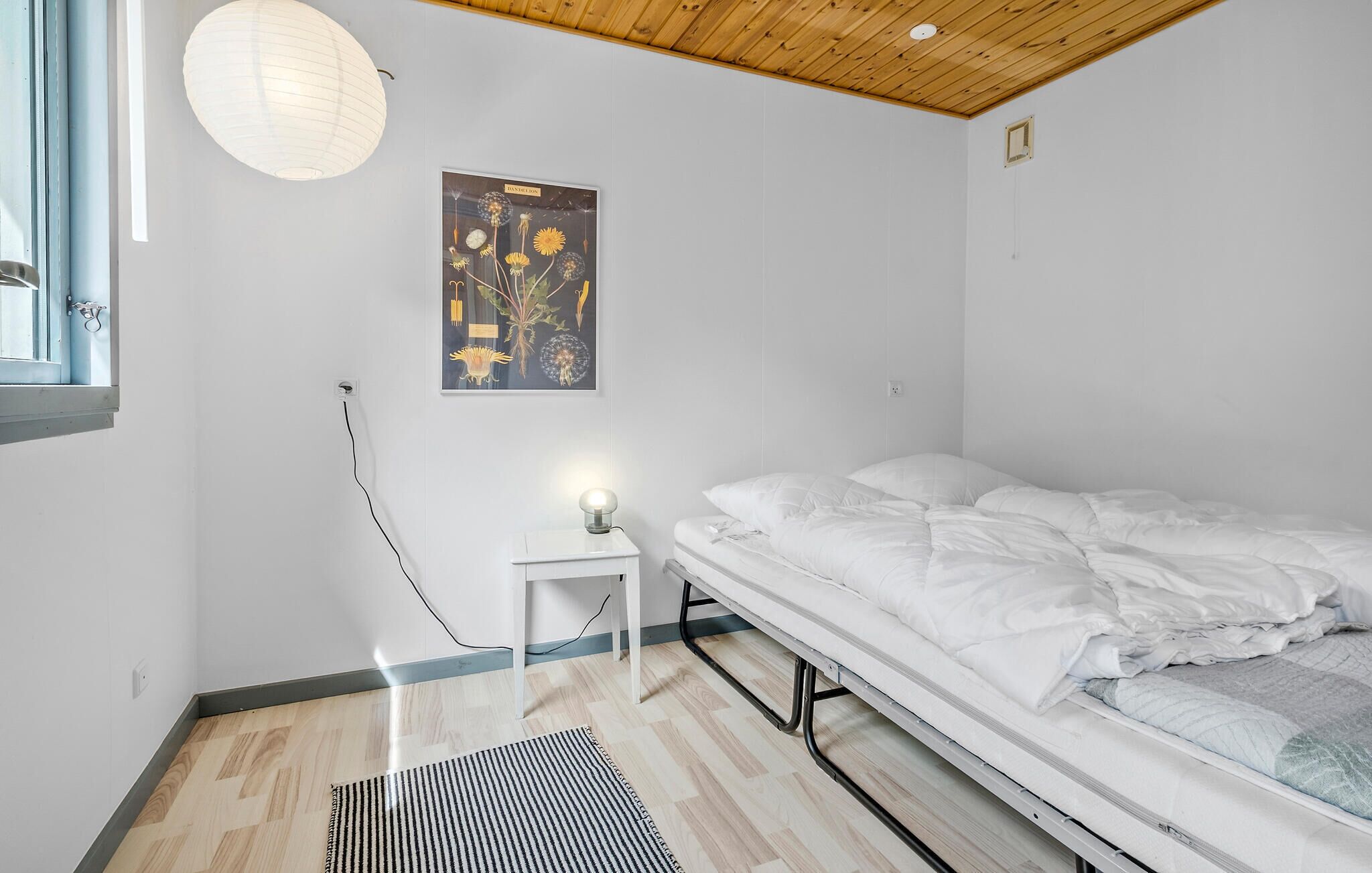 A bedroom with a bed, a small bedside table, a lamp, a wall-mounted picture, and a window with a curtain.