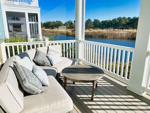 A covered outdoor seating area with a cushioned sofa and a round table, overlooking a body of water and a golf course.